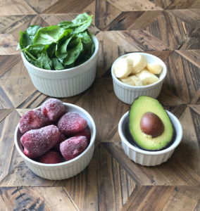 Spinach, frozen strawberries, banana and avocado in small bowls on a table.