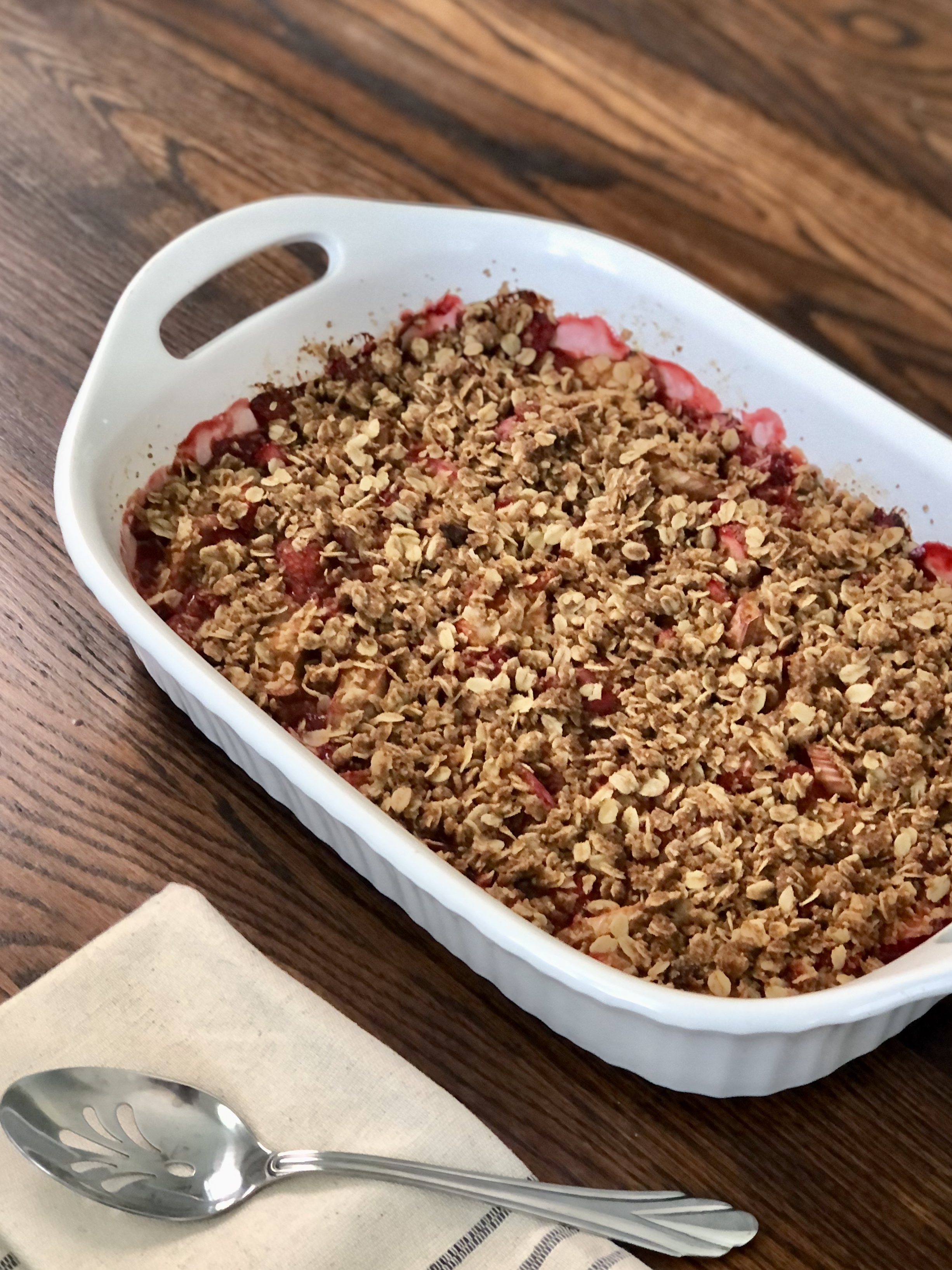 The baked strawberry rhubarb crumble in a white casserole dish on a wood table with serving spoon.