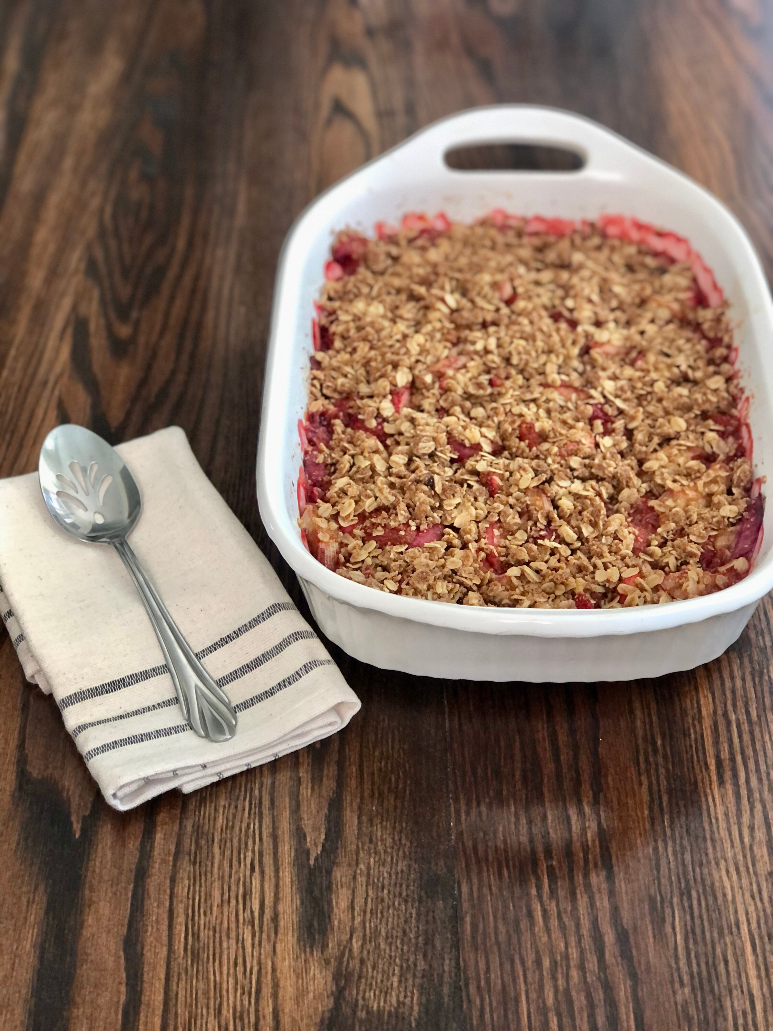 Baked Strawberry Rhubarb Crumble in white casserole dish on dark wood table with serving spoon on a cloth napkin.