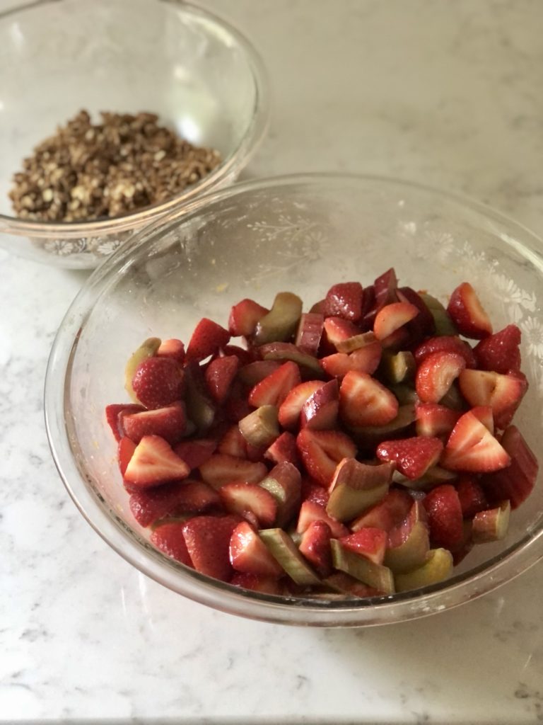Sliced strawberries and rhubarb in a large glass bowl and oat crumble in a small glass bowl on a counter.